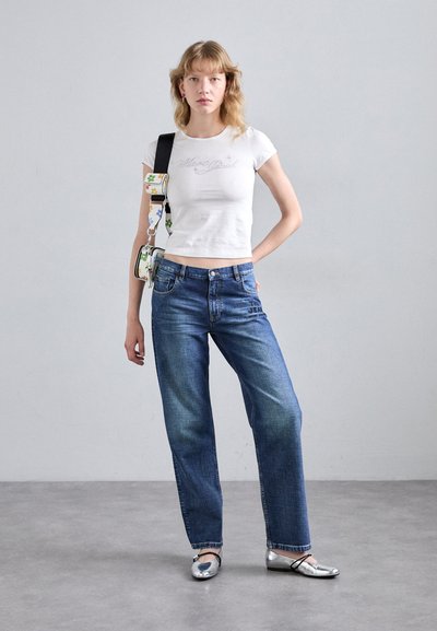 Giovane donna in piedi con una mano sui fianchi, indossa un top corto bianco, jeans blu, ballerine argentate e una borsa a spalla con motivi floreali.