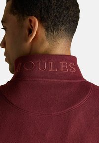 Bordeaux trui van een gestructureerde stof, met een geborduurd "JOULES" logo op de kraag en een hoge halslijn.