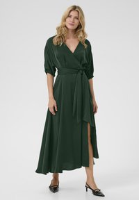 Vestito avvolgente verde scuro con maniche a sbuffo, vita con cintura e uno spacco laterale. Tessuto liscio con un design fluido. Indossato con tacchi neri a punta.