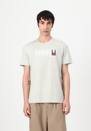 T-shirt en coton gris clair à manches courtes, col rond, arborant le logo "TOMMY" et un accent rayé en rouge, blanc et bleu.