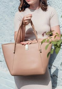 Bežová taška přes rameno s minimalistickým designem, hladkým veganským koženým materiálem, dvojitými držadly a kovovým akcentem na přední straně.