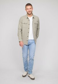 Chemise boutonée gris clair avec deux poches poitrine, portée sur un t-shirt blanc. Associée à un jean bleu clair et des baskets crème.