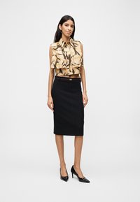 Γυναίκα που φοράει μπεζ μπλούζα χωρίς μανίκια με σχέδια, μαύρη στενή φούστα με ζώνη και μαύρα μυτερά τακούνια, στέκεται μπροστά σε απλό λευκό φόντο.