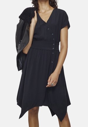 Žena oblečená v černých šatech na knoflíky s asymetrickým lemem, držící černou koženou bundu s beránkovým podšíváním přes jedno rameno.