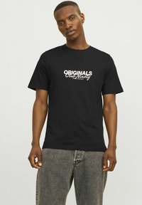 Camiseta de algodón negra con el texto en blanco y en negrita "ORIGINALS Club Winery" en el frente, cuello redondo y mangas cortas, combinada con jeans grises.