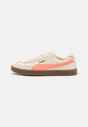 Niedriger cremefarbener Sneaker mit einem korallenfarbenen Streifen, brauner Sohle, cremefarbenen Schnürsenkeln und goldenem Puma-Logo an der Seite und Ferse.