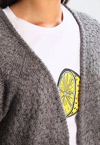 Harmaa pörröinen neuletakki, jossa avoin etuosa ja teksturoitu kangas. Alla valkoinen t-paita, jossa keltainen sitruunakuva.