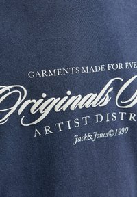 Námořnická modrá látka s bílým potiskem textu "Oděvy vyrobené pro každého Original Artist District Jack & Jones © 1990."