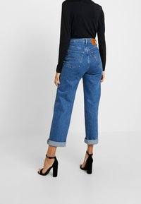 Person in blauem, hoch tailliertem, umgeschlagenem Jeans, schwarzem Langarmoberteil und schwarzen High-Heels, steht vor einem schlichten Hintergrund.