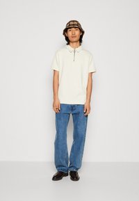 Cream polo shirt with a zip closure, paired with light blue straight-leg jeans and brown leather shoes. Bucket hat with a plaid pattern.