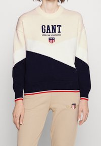 Pull en maille épaisse avec un design en blocs de couleurs crème et marine, poignets et ourlet côtelés, avec le logo GANT et un motif de blason sur la poitrine.