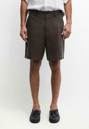 Mann trägt dunkelbraune knielange Shorts, ein in die Hose gestecktes weiß kariertes Hemd und schwarze Mokassinschuhe und steht vor einem schlichten Hintergrund.