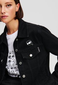 Črna denim jakna s srebrnimi деталji in belim grafičnim motivom na prsih. Obuta čez belo majico z enobarvnimi ilustracijami.