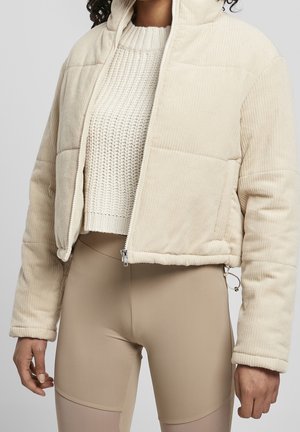 Donna che indossa una giacca di velluto a coste imbottita beige sopra un maglione bianco a maglia e leggings beige, in piedi contro uno sfondo semplice.