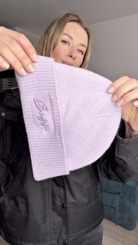 Λιλά πλεκτός σκούφος με πλεκτή μανσέτα και κεντημένο λογότυπο. Το ύφασμα φαίνεται απαλό, με ομαλή υφή και χωρίς ορατό σχέδιο.