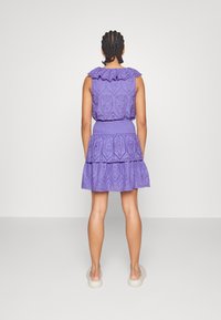 Donna con capelli trecciati che indossa un vestito senza maniche di pizzo viola con gonna a strati arricciata e scarpe bianche senza lacci, di spalle.