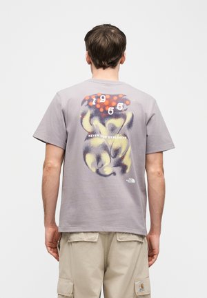 Man wearing light purple t-shirt with abstract dog graphic, red dots, "1996" and "Never Stop Exploring" text on back, paired with beige shorts.