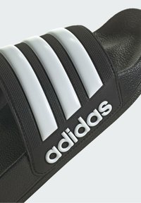 Svart Adidas sandaled med tre vita vertikala ränder, texturerad rem och upphöjd logotyp. Stängd baksida med platt sula.