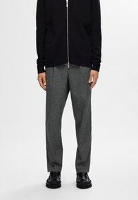 Schwarzer Strick-Cardigan mit Reißverschluss über grauen Karo-Hosen. Die Hosen haben einen lässigen Schnitt. Schwarze Schuhe mit strukturiertem Detail. Einfaches, modernes Design.