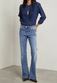Blusa blu navy con apertura a bottoni e parte anteriore testurizzata, abbinata a jeans chiari a zampa d'elefante e stivali marroni su un pavimento testurizzato.