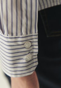 Poignet de chemise à manches longues rayée en blanc et bleu. Comprend deux boutons ronds et un ourlet cousu. Tissu doux avec une texture lisse.