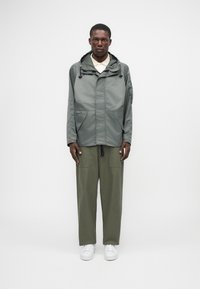 Ελαφρύ, μπουφάν με κουκούλα σε υποτονικό πράσινο, με φερμουάρ μπροστά, δύο μπροστινές τσέπες και ρυθμιζόμενα κορδόνια, συνδυασμένο με ελαιόχρωμες παντελόνες και λευκά αθλητικά παπούτσια.