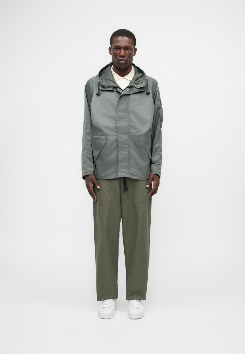 Ελαφρύ, μπουφάν με κουκούλα σε υποτονικό πράσινο, με φερμουάρ μπροστά, δύο μπροστινές τσέπες και ρυθμιζόμενα κορδόνια, συνδυασμένο με ελαιόχρωμες παντελόνες και λευκά αθλητικά παπούτσια.