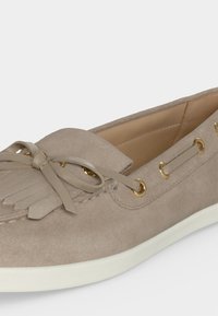 Mocassin en daim taupe avec nœud et détail à franges, œillets dorés et semelle en caoutchouc blanche, présenté en vue latérale inclinée.