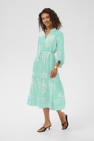 Tânără cu păr creț purtând o rochie midi verde mentă cu modele florale albe, brățară aurie și sandale negre cu toc, zâmbitoare.