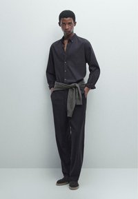 Camicia scura con maniche arrotolate, abbinata a pantaloni larghi scuri. Un maglione grigio legato in vita e scarpe scure.