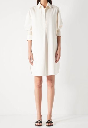 Robe chemise blanche avec un col à revers, des manches tombantes et des fentes latérales. Portée avec des sandales plates noires ornées d'accents dorés.