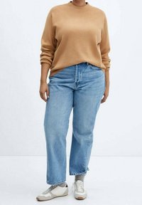 Suéter color camello, jeans de pierna recta azules y zapatillas blancas con detalles en gris. El suéter tiene un cuello redondo y mangas enrolladas.