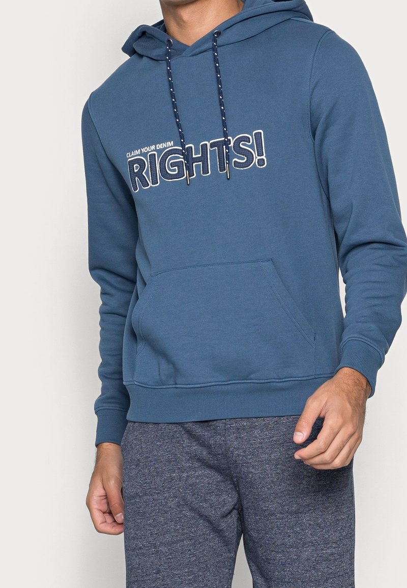 Homme portant un sweat à capuche bleu avec le texte « CLAIM YOUR DENIM RIGHTS ! » et des cordons assortis, associé à un pantalon de survêtement gris foncé.
