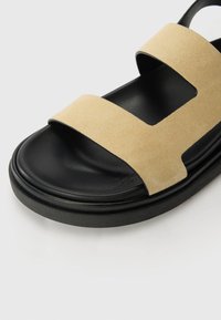 Sandales en daim beige avec deux larges brides et une semelle noire. Le design comporte des sections découpées pour la respirabilité. Simple et moderne.