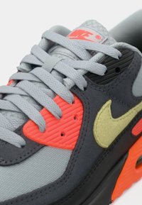 Sneaker Nike con tomaia grigia e nera, accenti arancioni neon, sovrapposizioni testurizzate e lacci grigi piatti con un logo distintivo.