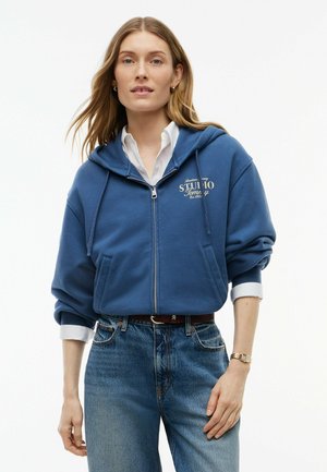 Kvinde iført blå lynlåshættetrøje med hætte, hvid skjorte med krave, blå jeans og ur på venstre håndled, stående mod hvid baggrund.
