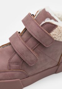 Chaussure en daim violet avec deux sangles Velcro réglables, doublure en fausse fourrure douce et semelle en caoutchouc épaisse avec des détails de couture contrastants.