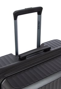 Valise rigide noire avec texture striée, poignée télescopique extensible et une poignée de transport fixe sur le dessus.