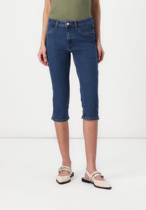 Frau trägt blaue, gekürzte Jeans und beige spitze Ballerinas mit Knöchelriemen, steht vor weißem Hintergrund.