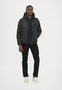 Schwarze Pufferjacke mit Kapuze, Reißverschluss vorne, Steppdesign und Logodetail. Kombiniert mit schwarzen Hosen und hellen Sneakers.