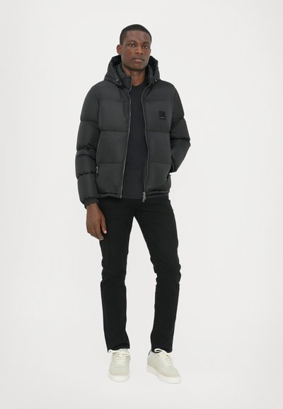 Doudoune noire avec capuche, fermeture éclair à l'avant, design matelassé et détail de logo. Associée à un pantalon noir et des baskets de couleur claire.