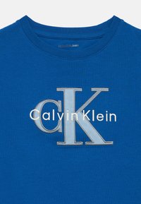 Zils kokvilnas T-krekls ar apaļu izgriezumu. Priekšpusē ir uzraksts “Calvin Klein” baltās un divtoņu pelēkās izšūtās burtiņās.