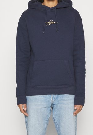 Persoon draagt een marineblauwe hoodie met een voorkantzak en gele "Hollister" tekst, gecombineerd met lichtblauwe spijkerbroek tegen een effen achtergrond.