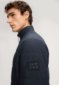 Granatowa kurtka puchowa z wysokim kołnierzem, o fakturowanym wykończeniu oraz naszywką z logo marki na lewym rękawie. Zamek przy szyi.