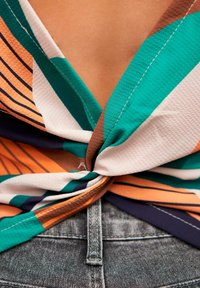 Blusa anudada en tejido multicolor con rayas y patrones abstractos, que presenta tonos de verde, naranja y marrón, combinada con una cinturilla gris oscuro.