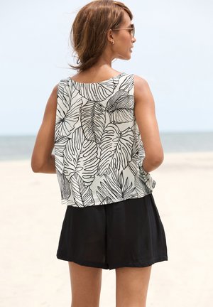 Leichtes ärmelloses Oberteil mit einem schwarzen Blättermuster auf einem cremefarbenen Hintergrund, kombiniert mit schwarzen Shorts. Glatte Stofftextur.