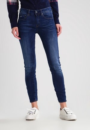 Personne portant un jean skinny bleu foncé et des baskets blanches, debout devant un fond clair uni.