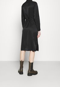Schwarzes Satin-Kleid mit tailliertem Gürtel und langen Ärmeln, mit seitlichen Schlitzen. Kombiniert mit dunkelbraunen Ankle Boots. Glatte Textur.