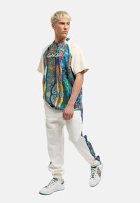 Kleurvol hemd met korte mouwen, versierd met abstracte patronen in blauw, groen en rood, gecombineerd met crèmekleurige joggers en witte sneakers met accenten.