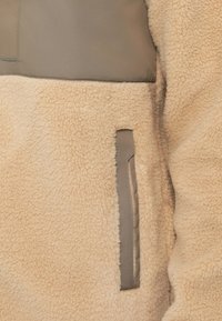 Beige Fleecejacke mit einer taupefarbenen Stoffbrusttasche und einer mit taupefarbener Borte versehenen Reißverschlusstasche auf der Vorderseite.